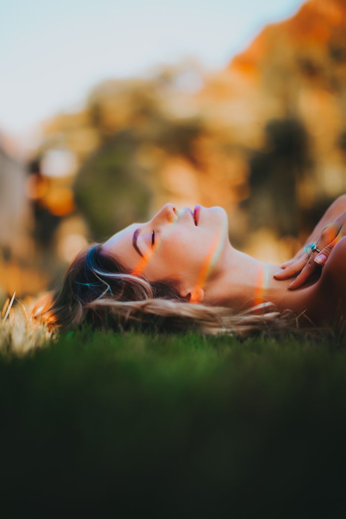 services-01 A woman with closed eyes lies peacefully on grass, bathed in warm sunlight.