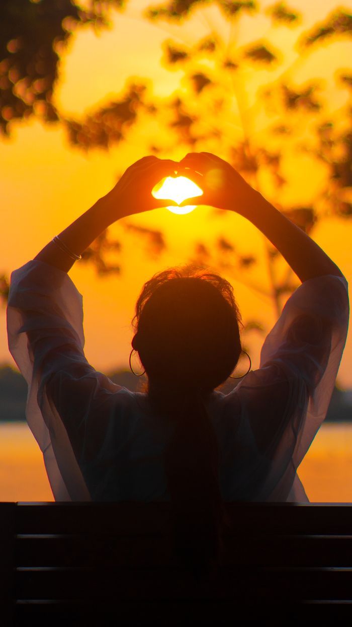 ours-journey Backlit silhouette of a woman forming a heart shape with her hands against a stunning sunset.