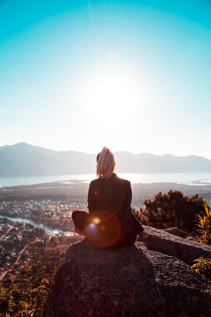 contact-img A woman sits on a cliff, gazing at a stunning valley landscape under a bright sun.